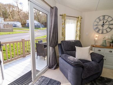 A living room with a large armchair a sideboard with a clock on the wall and sliding glass doors leading to a patio at 98 The Heron in New Quay