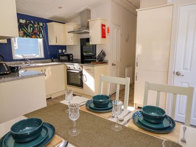A kitchen with cream cabinets a built-in oven and stovetop a dining table set with blue plates bowls and glassware at 98 The Heron in New Quay