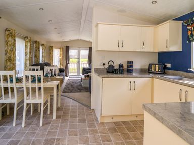 A kitchen with cream cabinets and gray countertops next to a dining area with white chairs and a living room with sofas at 98 The Heron in New Quay