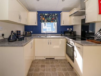 A kitchen with cream cabinets grey countertops a window with a floral blind and various appliances at 98 The Heron in New Quay