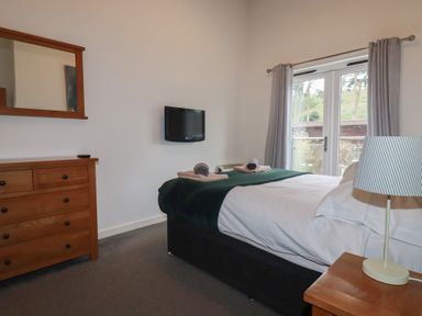 A bedroom with a bed a wooden dresser a wall mirror a wall-mounted tv and a lamp on a bedside table at Treburgett in Porthtowan