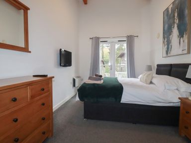 A bedroom with a bed a wooden dresser a wall-mounted TV and double doors with curtains at Treburgett in Porthtowan