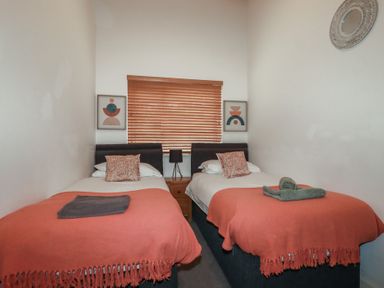 A bedroom with two single beds with orange throws and pillows a window with wooden blinds and framed abstract art on walls at Treburgett in Porthtowan