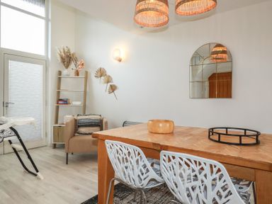 A dining area with a wooden table and white chairs next to a beige armchair and ladder shelf at Trenethick in Porthtowan