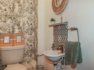 A small bathroom with a toilet, a sink with a green towel, patterned tiles behind the sink, and decorative items on the toilet and a shelf at Trenethick in Porthtowan