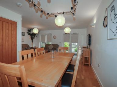 A dining table with wooden chairs in a room with sofas plants and wall art at Tregullan in Porthtowan