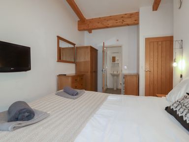 A bedroom with a bed towels on bed wooden wardrobe dresser wall mirror wall mounted tv and an open door to a bathroom at Tregullan in Porthtowan