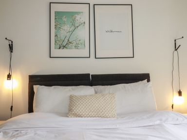 A bed with white bedding and pillows with two framed pictures above and two hanging light bulbs at Treveglos in Porthtowan