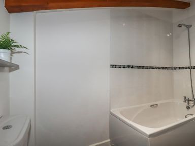 A bathroom with a white bathtub showerhead toilet wooden ceiling beam and a plant on a wall shelf at Treveglos in Porthtowan