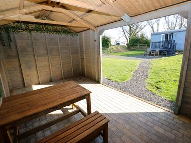 A wooden gazebo with picnic benches overlooking a grassy yard with a path leading to a small cabin and outdoor seating at Little Acre Oxenhope