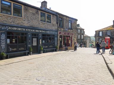 A street with shops including Haworth Steam Brewing Co and Pave Cheesemonger with people walking and a red phone booth in Haworth