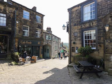 A cobblestone street with Cafe Verde and Black Bull buildings and outdoor seating at Little Acre in Oxenhope