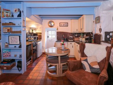 A kitchen with a round dining table flowers on table brown armchair with cushions blue shelving unit and white cabinets at Llety in Beddgelert