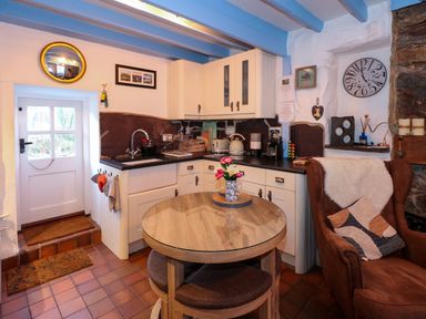 A small kitchen with white cabinets a round wooden table with flowers a brown armchair and a door at Llety in Beddgelert