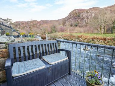 A bench with cushions on a deck overlooking a river and hills at Llety in Beddgelert