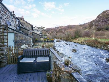 A riverside stone terrace with a bench and cushions beside a flowing river at Llety in Beddgelert