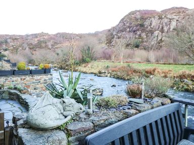 A stone dragon sculpture on a rock wall by a river with plants and a bench at Llety in Beddgelert