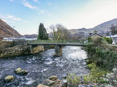A river flowing under a small stone and metal bridge with houses and hills in the background at Llety in Beddgelert