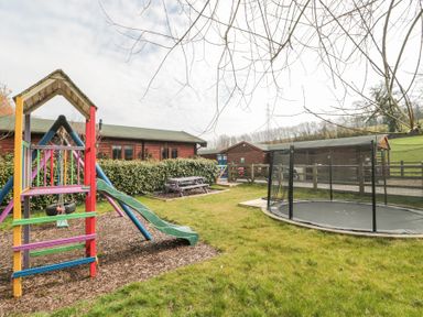 A garden with a colorful slide and swings set a trampoline a purple picnic table and wooden buildings at Bell tent 2 in Camerton near Timsbury