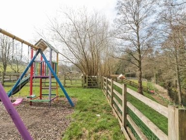 A playground with swings and slides next to a fenced grassy area with trees and a tent in the distance at Bell tent 2 Camerton near Timsbury