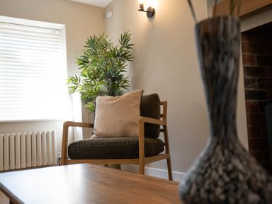 A wooden armchair with cushions near a window with blinds and a green plant at Driftwood in Kessingland
