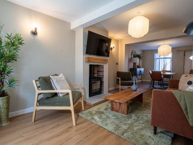 A living room with two armchairs a wooden coffee table a green rug a wall-mounted tv and a dining area at Driftwood in Kessingland