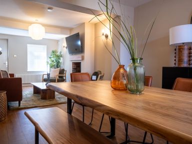 A dining area with wooden table and bench with vases a living room with chairs sofa coffee table and wall mounted TV at Driftwood in Kessingland