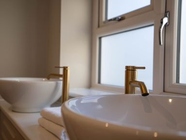 Two white vessel sinks with gold faucets and folded towels in a bathroom with a window at Driftwood in Kessingland