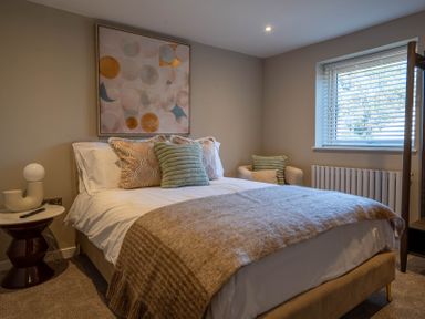 A bedroom with a bed with pillows and a blanket a side table with a lamp an armchair and a window at Driftwood in Kessingland