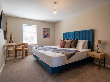 A bedroom with a blue upholstered bed with pillows a wooden bedside table with a lamp a wooden desk with two vases a brown chair a window with blinds and a wall painting at Driftwood in Kessingland