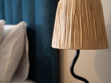 A close-up of a beige lampshade with a black curved lamp base next to a bed with white pillows and a dark blue headboard