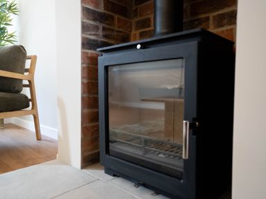 A black wood stove set in a brick fireplace next to a wooden armchair with green cushions
