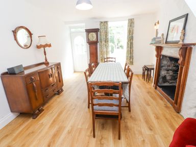 A dining room with a wooden table covered by a tablecloth surrounded by six chairs next to a mantelpiece with decorations and a sideboard with a lamp and mirror at Llys Emrys in Beddgelert