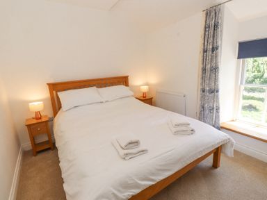 A bedroom with a wooden bed frame white bedding bedside tables with lamps and a window with patterned curtains at Llys Emrys in Beddgelert