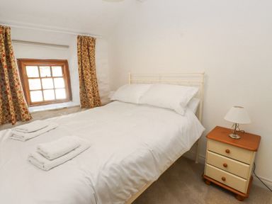 A bedroom with a double bed white bedding bedside table with lamp and window with floral curtains at Llys Emrys in Beddgelert