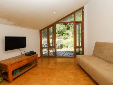 A room with a beige sofa a wooden media console with electronics a wall-mounted TV and a glass door leading to a garden at Llys Emrys in Beddgelert