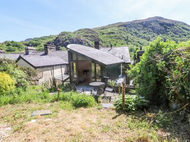 A garden with patio chairs and table outside a house with sloping roof and hills in the background at Llys Emrys in Beddgelert