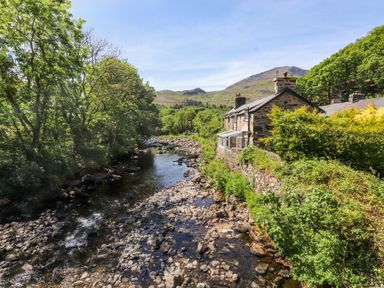 A river with rocks bordered by trees and a stone house near hills at Llys Emrys in Beddgelert