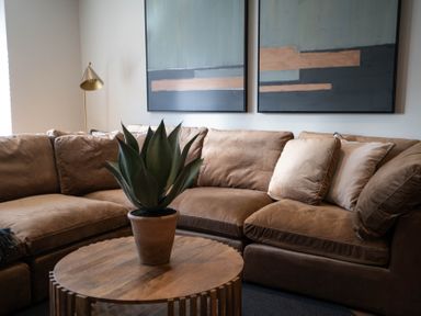A brown sectional sofa with cushions a wooden coffee table with a potted plant and two abstract paintings on the wall at The Reef in Kessingland
