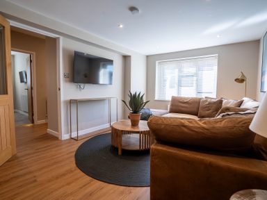 A living room with a wall-mounted TV a round wooden coffee table with a plant on a black circular rug and a brown sectional sofa at The Reef in Kessingland