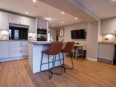 A kitchen with an island and two brown chairs a dining table with chairs and a TV on the wall at The Reef in Kessingland