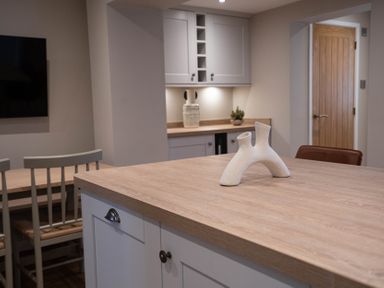 A kitchen with a wooden countertop island a white abstract vase chairs a wooden table cupboards and a wooden door at The Reef in Kessingland