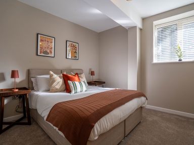 A bedroom with a bed with pillows and blanket two bedside tables with lamps two framed pictures and a window with blinds at The Reef in Kessingland