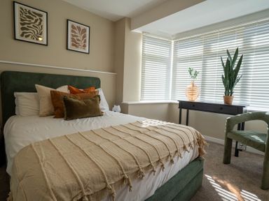 A bedroom with a bed green headboard pillows and a beige blanket with a black side table and green chair near windows with blinds at The Reef in Kessingland
