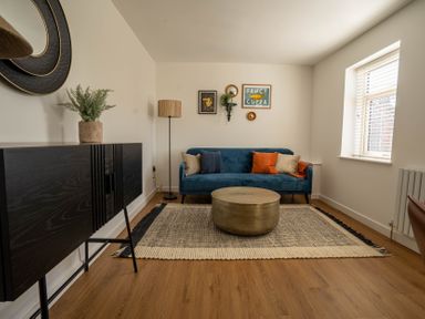 A living room with a blue sofa cushions round coffee table black cabinet wall art and a window at The Cove in Kessingland