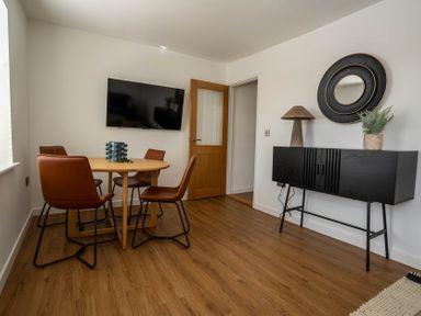 A dining area with a round wooden table and four brown chairs a wall mounted flat screen tv a wooden door a black sideboard with a lamp plant and circular mirror on the wall at The Cove in Kessingland
