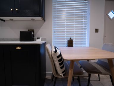 A kitchen corner with black cabinets a wooden table chairs with cushions and a window with blinds at The Cove in Kessingland