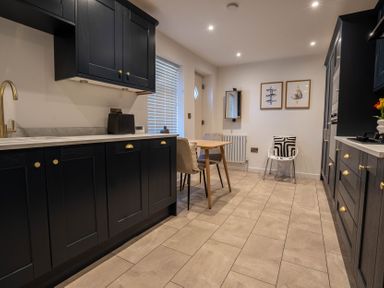A kitchen with black cabinets a wooden table with chairs a white chair with a black and white cushion and framed pictures on the wall at The Cove in Kessingland