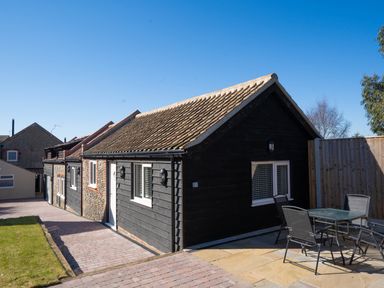 A black wooden house with a tiled roof and outdoor table with chairs in a paved yard at The Cove in Kessingland