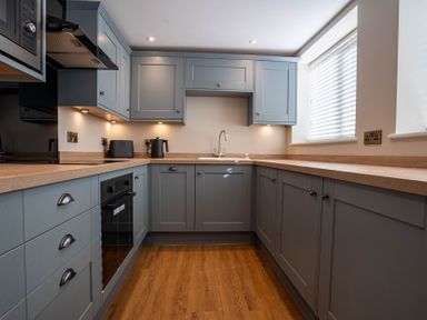 A kitchen with gray cabinets wooden countertops an oven a sink a kettle and a window with blinds at Coral Cottage in Kessingland